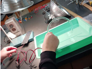 A lamp shines above a solar module which is wired to a device in a green basin of water, unseen by a hand. A clear tube is held in the hand and water is coming through the tube.