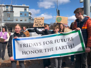 Students hold a banner that reads, "FRIDAYS FOR FUTURE HONOR THE EARTH!" More students in the background, one holds cardboard signs that read, "Climate Action Now".