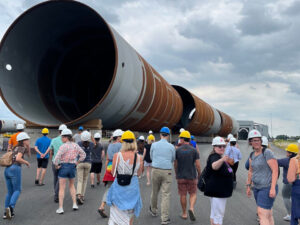 Group of people in hard hats approach large rusting metal cylinders of a retired wind turbine