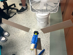 Kidwind wind turbine model with three rectangle blades sitting on the floor in front of a box fan sitting on a bucket.