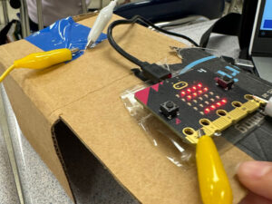 Microcontroller board connected with alligator clips to the top of a cardboard box near blue tape. USB cable attached to board.