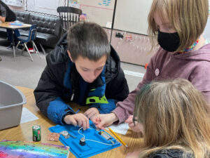 3 kids work at a blue plastic tray with a C-cell battery and two wires connected to a motor.