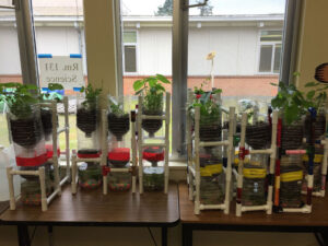8 Pipe constructed towers hold a large plastic bottle with soil and a plant. Draining into a lower plastic container with soil or pebbles and a 3rd container with water and colored beads.