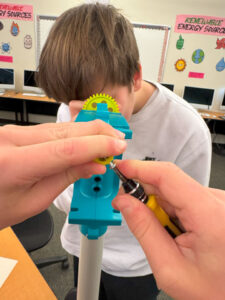 Two hands hold a screwdriver and are inserting a screw into a yellow circle that is obscured by the hands. It is attached to a teal mounting apparatus. On the far side of the device another student with short brown hair and a white shirt looks closely at a yellow gear.