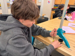 A student uses a tool to make an adjustment to a yellow item with a dowel sticking out of it. Next to the item is the base of a KidWind turbine.