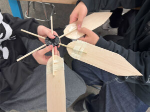 A top down photo of two students facing each other sitting in chairs. They are both holding a black plastic hub with 6 dowel. 2 blades have been attached on the near and near right dowel with masking tape. One student attaches a third blade with packing tape.