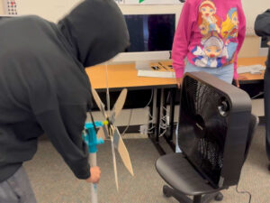 A student in a black hoodie braces a KidWind turbine in front of a black box fan sitting on a chair. The turbine has 6 hand cut wooden blades taped to dowels.