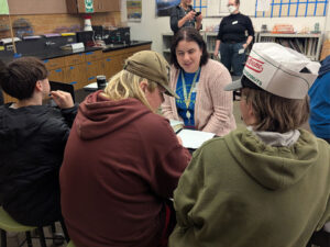 A woman with a blue shirt, lanyard, and pink cardigan sits at a table. Three students face her.