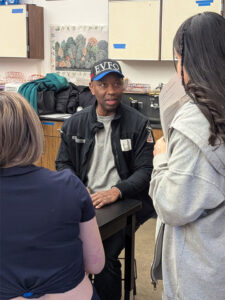 A man wearing a black hat that says EVFO and a black jacket with patches sits at a black classroom table facing the camera. Two girls with their backs to the camera look at him. The girl on the left is blond with a blue shirt, the girl on the right has dark hair and a grey hoodie. She is holding a paper near her mouth.