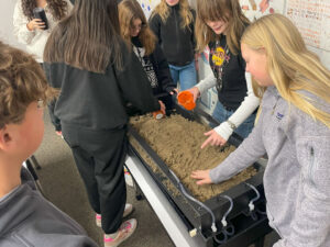 A group of girls surround a water table filled with tan sand. Two girls hold orange cups. The water table has three tubes connected on the near side.