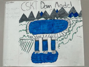 Photo of a white paper with a drawing of a dam. The drawing is labeled CSKT Dam Model. It has a row of mountains that are black peaks with a few blue peaks mixed in. In front of the mountains is a top-down view of a water basin in an oval shape with steep walls. To the bottom is a dam with three water channels and some frothy water in a lower oval of water.