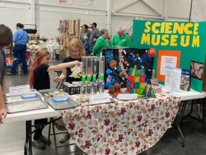 Photo of a table at a community event. On the table is a green tri-fold poster that reads Science Museum in large yellow letters. There is an orange square with a white paper in the middle that reads "Lets Build a SCIENCE MUSEUM. It is partially obstructed by a standing folder flyer that can't be read in the photo. To the left on the table is a model of the solar system, magnetic building toys, 4 pairs of connected litter bottles labled Tornado Simulation. Behind that cups with dirt can be seen. The leftmost part of the table has 4 trays one with markers and two with Tupperware containers and all trays have clear plastic pipettes.