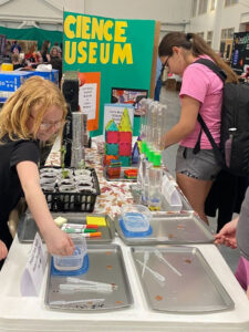 A table setup at a community event. A kid puts their hand in a Tupperware container 1/8th filled with a clear liquid. The container is on a grey tray, one of 4. The trays have clear pipettes. One tray has markers and sticky notes instead. Plants growing in white cups are in a black basket. Liter bottles connected with a green connector are standing behind the trays on the right. Magnetic building toys are behind the bottles and the A woman in a pink shirt with a black backpack is looking at something on the table by the magnet toys. At the far end of the table is a green tri-fold poster the large yellow letters: "CIENCE" and "USEUM" can be seen