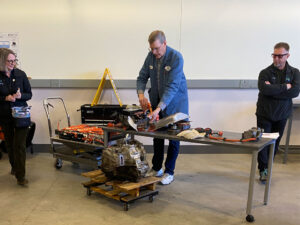 Three adults stand by a metal table on wheels. On the table is an electric component with batteries and a large power cable. A metal cover, a large metal coil a pair of gloves and a power cable attached to an unidentified device. In front of the table a motor sits on a pallet cart. To the left of the table is an array of batteries connected with orange cables.