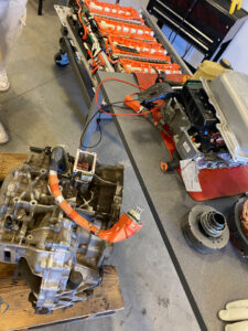 Top down photo of a table and 2 cards. On the table is a metal device with wires coming out, it is mostly covered by a metal lid. The cart to the left has a motor with a large electric cable coming out of the top. On the cart at the top orange power cables connecting what is likely batteries can be seen.