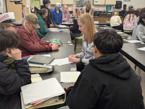 A woman sits at a black classroom table speaking to 4 boys. On the table are binders, papers, and a closed chromebook. In the background more groups of students are gathered around adults at connected black tables.