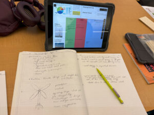 A student notebook sits open in front of a propped up tablet. On the tablet is a pie chart, notes, and a pros/cons chart. To the left of the chart is a picture labeled Offshore Wind Power, and part of picture labeled Land-based wind power. In the notebook is student writing and a drawing of a wind turbine with 4 blades. A yellow mechanical pencil rests on the notebook.