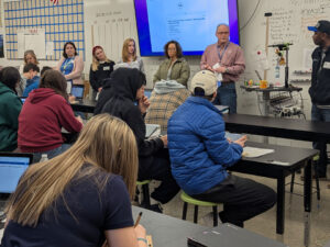7 Adults stand in front of a classroom. The students sit at black classroom tables with Chromebooks or paper and pencils facing the adults. On the Screen is a document with the PGE logo on it.