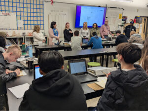 7 adults face a classroom of students sitting at black tables. Students are taking notes in a notebook or on Chromebooks. The adults are looking towards a woman in the middle.