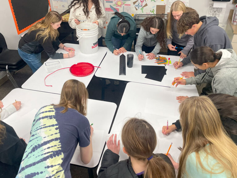 Four white topped work tables are pushed together with a small square of space in the middle. Students surround the tables with their heads down, all are holding a pencil. They have white paper on the table top. There are additional pencils, black paper, a mug, and a bucket on the top most tables.