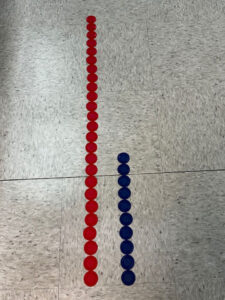 A photo of poker chips on the floor. A red line of chips extends one chip longer than three floor tiles on the left. On the right, blue chips are lined up across one tile and two chips onto the second.