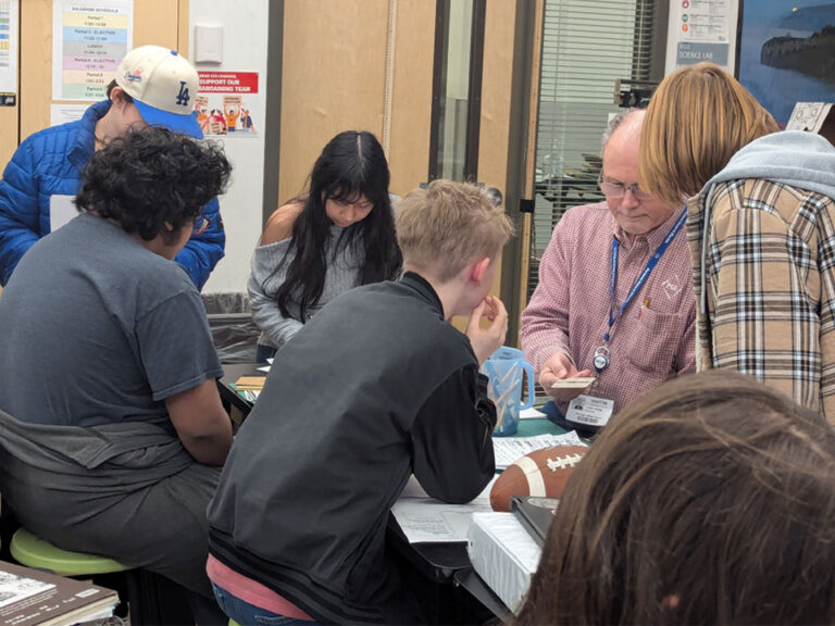 A man shows a card to a girl next to him while 4 students listen in from across a black classroom table. On the table a blue jug of pencils can be seen. In the foreground on the table is a binder and closed chromebook next to a football.