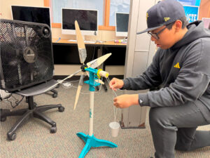 A student in a black hoodie and ball cap with yellow accents kneels on the floor next to a KidWind turbine. He is adjusting strings attached to a paper cup and the turbine via a spool. The wind turbine has 5 hand-cut blades taped to dowels and is sitting on the floor in front of a black box fan resting on a rolling chair.