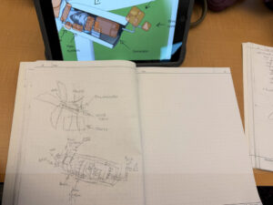 A sketch on graphing paper in a notebook shows the detailed components of a wind turbine exterior and interior. Labeled parts that can be read are: Hub, Nacelle, Anemometer, Wind Vane, Tower, Main Shaft, Pitch System, Hub, Rotar, Brake, Yaw System. In the background is a tablet with a color cutt-away of a wind turbine with labels. The viewable labels are: Wind vane, Generator, Yaw system.