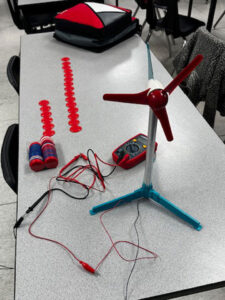 On a grey classroom table sits a wind turbine model with a single piece red 3 blade front. Behind it is a multimeter and some poker chips. Behind the tray of poker chips red chips are laid out in two rows on the table. The row on the right is twice as long as the row on the left.