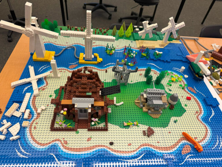 A LEGO model. On an island printed set of baseplates is a brown LEGO house with a black LEGO plate mounted near the roof. A grey tower and a grey and brown bunker building are also on the island. In the water are two wind trubines and sea plants. Across the water is another island with plants and two smaller wind turbines.