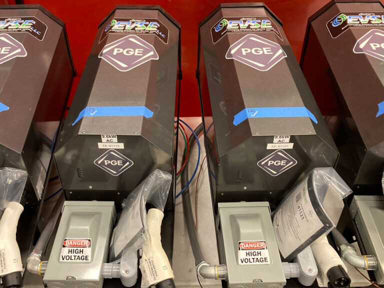 A row of 4 EV pole chargers. A black rectangular box with a half hexagon top side. At the top it reads EVSE below that is the PGE logo. At the bottom is a strip of blue tape with the number and a checkmark. Below the box is a connected grey box labeled Danger High Voltage and an EV charging port.