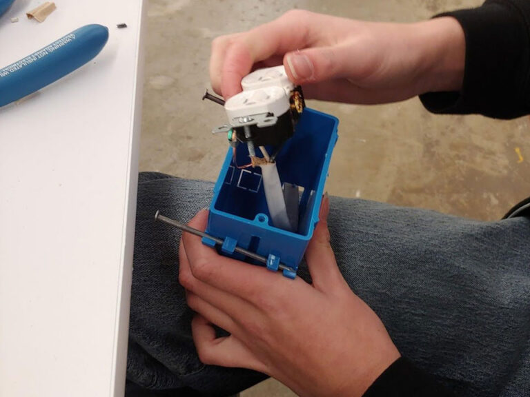 A blue el;etrical gang box is having a white electrical outlet inserted from the top. The gang box is being held in a pair of hands. Wires can be seen entering the center of the box.