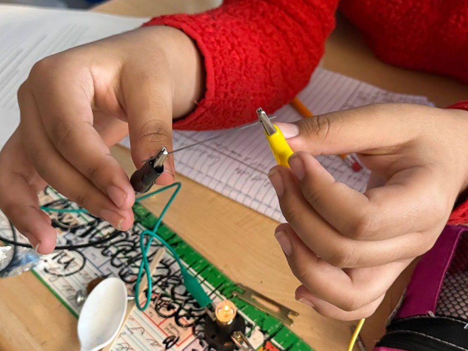 Two hands each hold an alligator clip test lead. The clips are attached to a graphite pencil lead. On the table below the hands, the other end of the test leads connect to a lit E10 light, the rest of the circuit can not be seen.