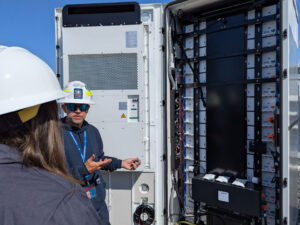 View over the shoulder of someowe with long brown hair under a white hard hat. A man stands before an open battery storage container both hands point towards the array of batteries inside the box.