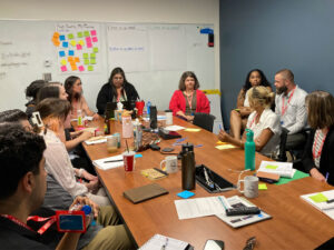 Educators and industry experts gather around a table covered in notepads. Behind them is a white board with mulit-colored sticky notes on a paper that reads "High Quality PD/ Training"