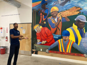 A man in a blue polo stands tot he left of a mural. The mural shows 5 people in safety gear before green hills. One person is using a tool on a wooden structure. Anoter carries wooden planks that read "Work Ethic, Skills, Persistence, Assertiveness, Physical & Mental Toughness, Life Outside of Work, Choices" Another person uses an angle grinder on a metal box, A man looks at a blue print and at the bottom a man holds a red level that reads "Leveling the playing field"