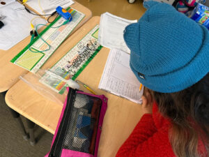 A student in a blue knit hat leans over their desk using a pencel to write into a notebook. The notebook is turned sideways and has three columns, writing can not be read. On the desk in front of the student is a bag with a plastic spoon, another penciles and a small screw. On the next desk in front of the student is a blue D-Cell battery holder with battery a green alligator test lead connects to a light bulp. A yellow test lead runs off the top of the image and a black test lead returns to the battery pack.