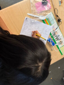 A top down view of a student's head. Slightly blurred in the background is a notbook with writing written perpendicular to the lines. A plastic bag with a plastic spoon some pink sticky notes a blub with a black and a white test lead connected and a blue D-cell battery holder.