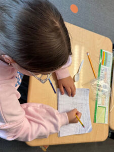 A student fills in a notebook page that is turned sideways and has three columns drawn. Each column is half filled with handwriting.