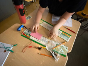 The image looks down on two student desks, the near desk has a name bar that reads Owen. Over the center where the two desks meet a student holds a yellow test lead and a black test lead. The test leads are pressed into an item under their right hand that may be aluminum foil resting on a blank pink sticky note. The yellow test lead is connect to a lit light. A red test lead connects the light to a blue D-cell battery pack and battery which is connected to the black test lead held by the student. A plastic bag with a plastic spoon a pencil and other items that can nobe be clearly identified rest on the desk by the light.