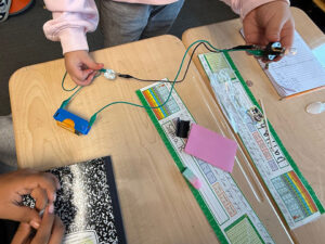 A student stands behind two pushed together desks . In one hand they hold a light bulb that is lightly glowing. A black and a green test lead run down from the light. The black wire connects to one side of a square of aluminum foil. The student holds a green wire to the other side of the aluminum foil. Both green wires connect to a blue D-cell battery holder with an orange D-cell battery.