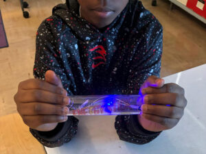 A middle school student holds a Steve Spangler energy stick between his hands. Each hand fully caps an end of the energy stick. The energy stick is glowing blue in the middle which is also reflected on the inner surface and between the fingers of the left hand.