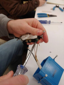 A student uses a screwdriver to connect a grounding wire to an electrical outlet. The white and black wires have been hooked and wait below the outlet with the wire pulled through a blue gang box. More hand tools can be seen in the background on the white work table.