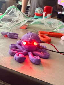 On a desk with open zip lock bags and scissors is a puple clay sculpture shaped like an octopus. It has a red and a black whire connected to the clay on the right side. Two red lights are lit making eyes in the middle of the head. There is a lighter colored band of clay behind the eyes that bisects the sculpture.