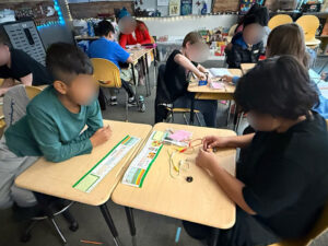 Students with their faces blurred sit at classroom desks in pairs of twos. The desks in the middle of each pair. On the desk in the foreground the student on the righ manipulates an item with alligator test leads and a light and more materials in a plastic bag. Across the desks to the left the other student leans on his desk to watch.
