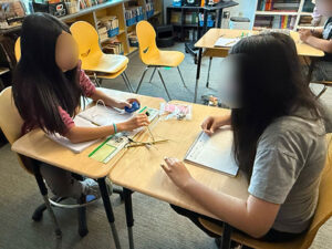 Two students with their faces blured sit at desks that have been pushed together to face each other. Each student has a notebook on their desk. The student on the left is holding a light connected to a blue plastic item in her other hand.