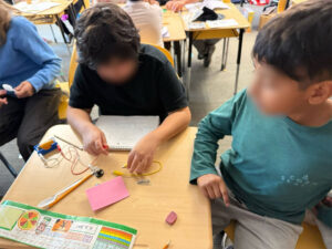 Two students share a desk, their faces blurred. On the desk is a plastic spoon, a pencil, a blank pink sticky note, 2 screws, a blue D-Cell battery holder with battery. Connected to the battery holder are a red and a white whire. The student behind the desk is holding the red wire. A yellow wire lays under his left hand on the desk by the screws.
