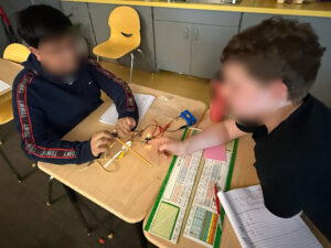 2 students are seated at desks that have been pushed together to face each other. The student on the righ leans over a notebook on the desk to point at a lit light on the other desk. The student on the left is holding two wires to a yellow pencil near the eraser. The wires are black and white and connected to a blue D-cell battery holder.