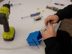 A student is stripping the wire wire of a household wiring cable (Romex) which is pulled through a blue gang box and continues across the white worktable to the left. On the table is an electric drill two screwdivers an electric outlet three yellow wire caps and a clipboard with a pen.