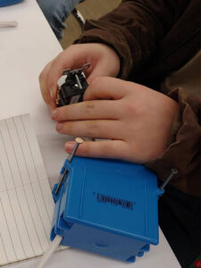 A student holds an electric outlet with the back facing the camera. Under their left hand a gang box with a white electric cable rests on the white worktable.